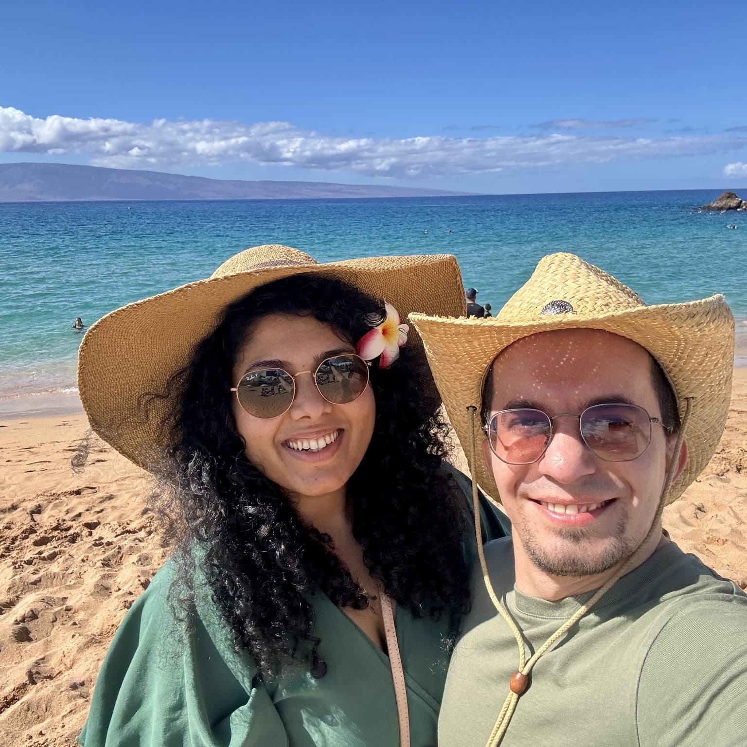 Gayatri and Oleg in straw hats taking a selfie on a sunny beach with turquoise water behind them