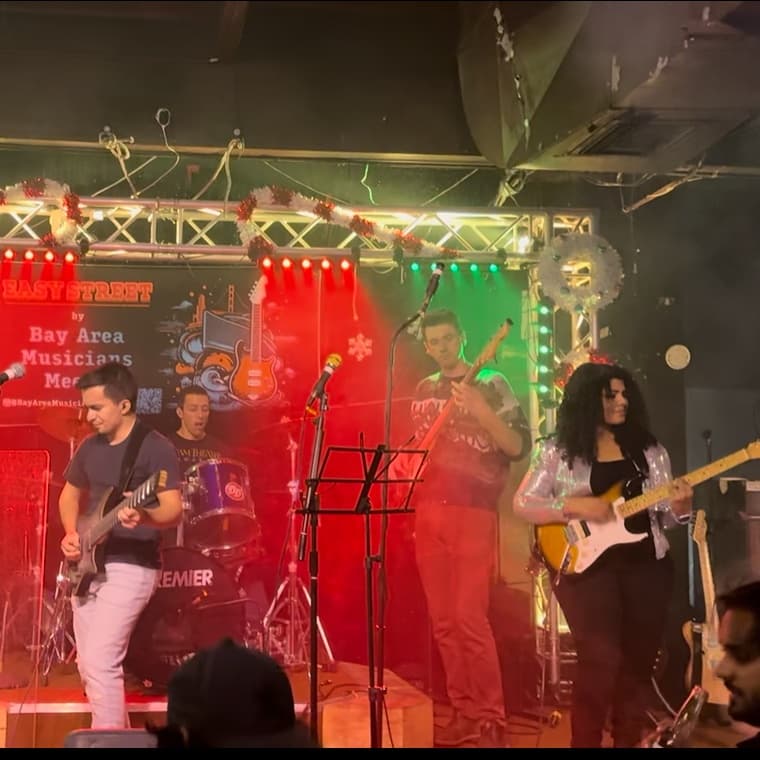 Gayatri and Oleg playing guitar with a band on the holiday-lit Easy Street stage at Bay Area Musicians Meetup